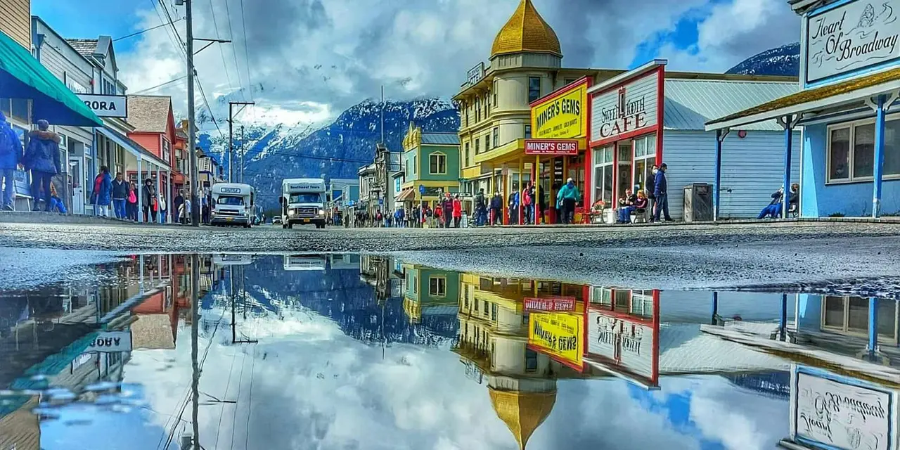 Colorful wooden storefronts on Skagway's main street with snowy mountains behind, reflected in a puddle
