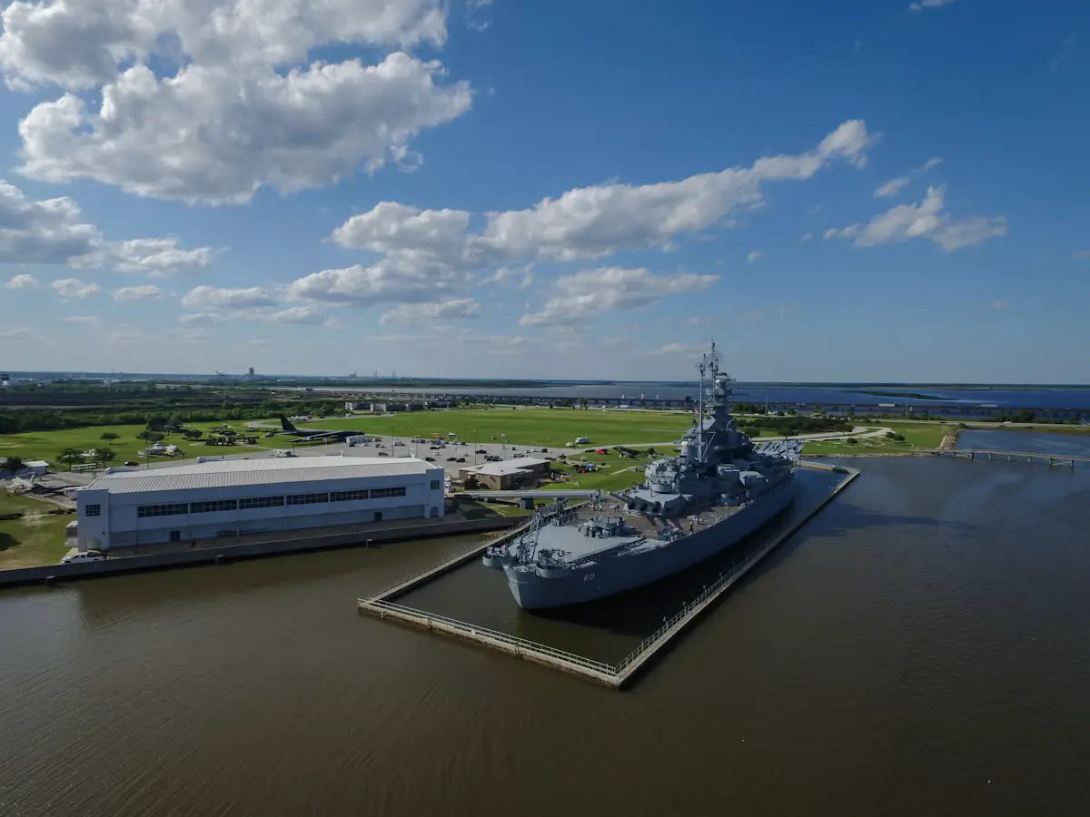 Aerial view of the battleship USS Alabama moored on Mobile Bay at Battleship Memorial Park, under a blue sky with big white clouds