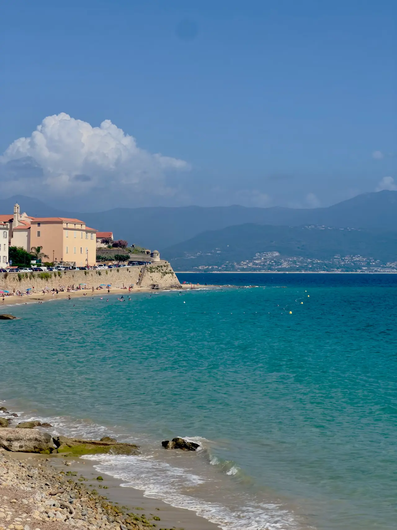 Turquoise water and a pebble shore at Plage Saint-François, with ochre houses on the old sea wall at left and green mountains across the Gulf of Ajaccio under a blue sky