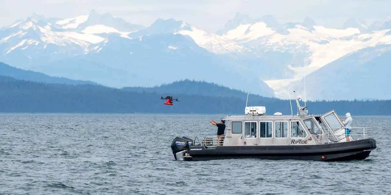 A small tour boat on grey-blue water with a crew member launching a drone beside it and snow-capped mountains behind
