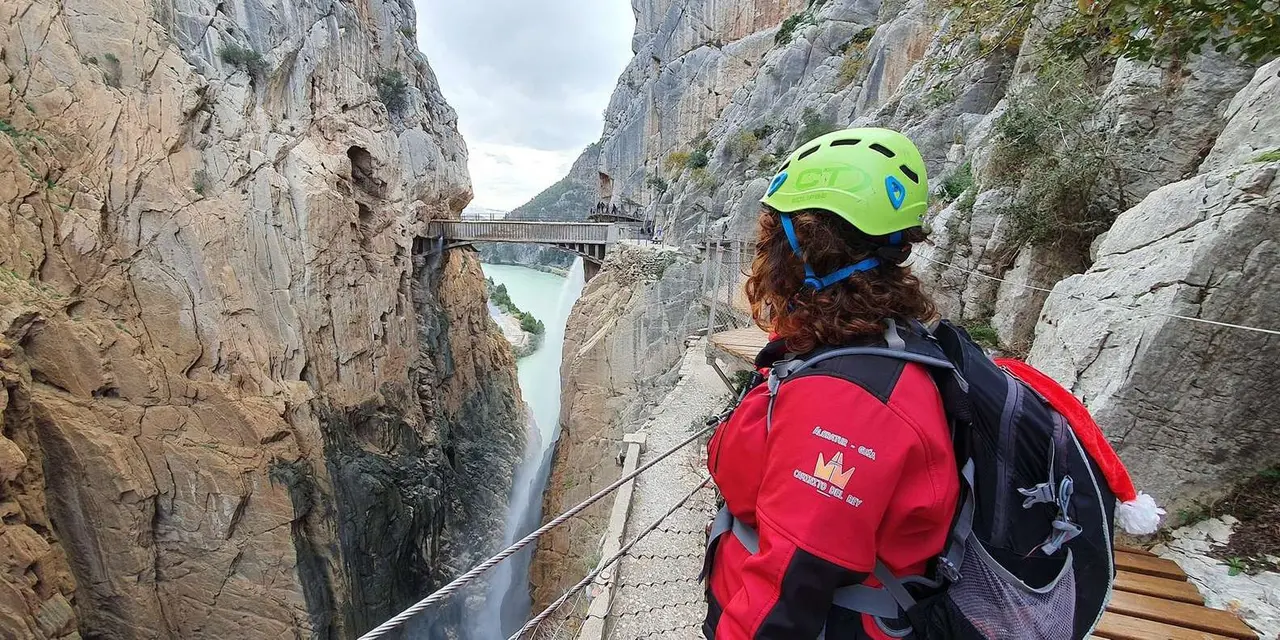 A hiker in a green helmet and red jacket on a narrow cliffside walkway high above a gorge, with a waterfall and a bridge in the background