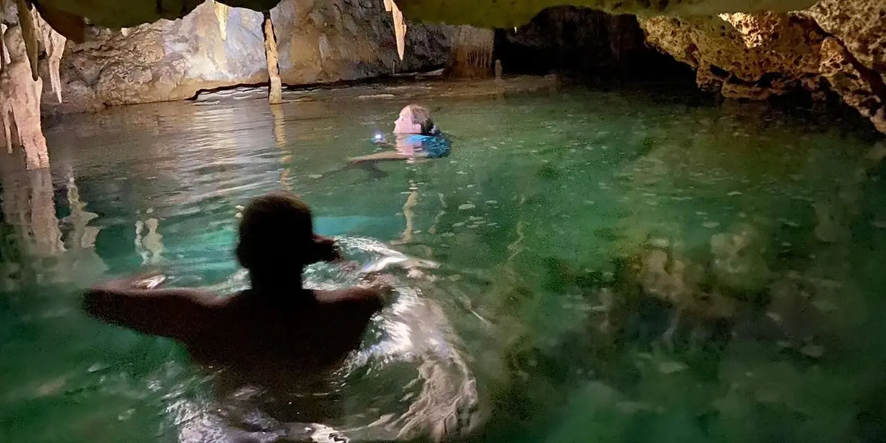 A person swimming in a clear green pool inside a cave beneath rock walls