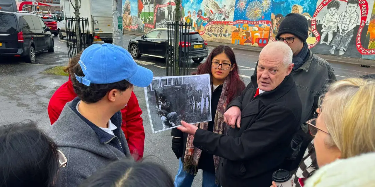A guide holding up a black-and-white photograph to a group of visitors on a street, with a long mural wall behind them