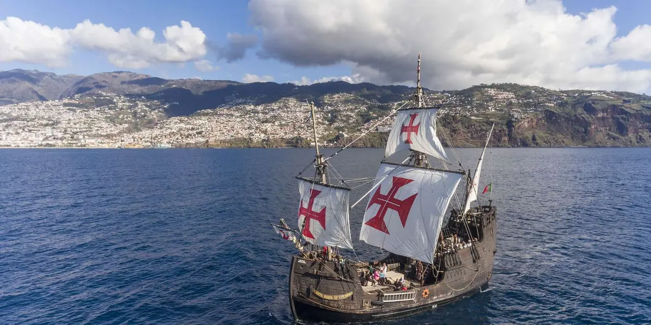 A wooden replica ship with red-cross sails on open water, the hillside of Funchal rising behind it