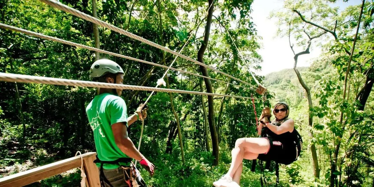 A zip-line guide in a green shirt stands on a wooden platform while a woman in a helmet and harness glides along the line through jungle trees