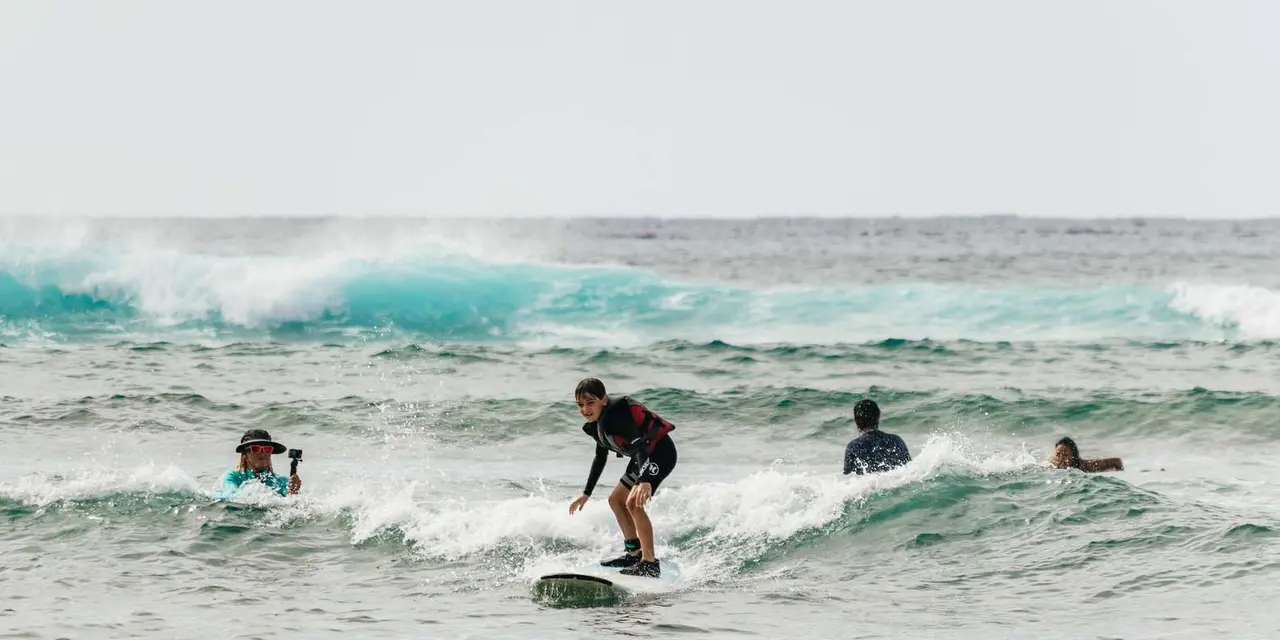 A surfer standing up on a small breaking wave off Waikiki while another student paddles behind