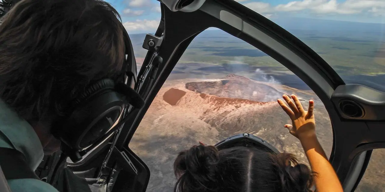 A child's hand pressed against a helicopter window above a brown volcanic crater