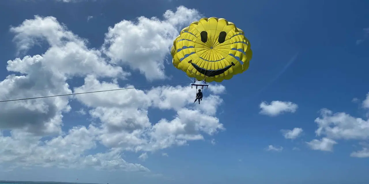 A yellow smiley-face parasail with a rider hanging below it, high over blue sea under a sky of white clouds