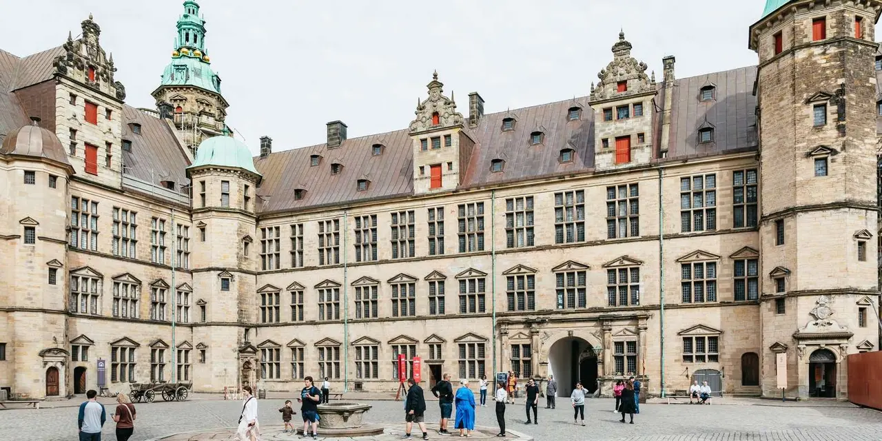 A sandstone castle courtyard with green copper-roofed towers and visitors walking across the cobbles in front of the arched gateway