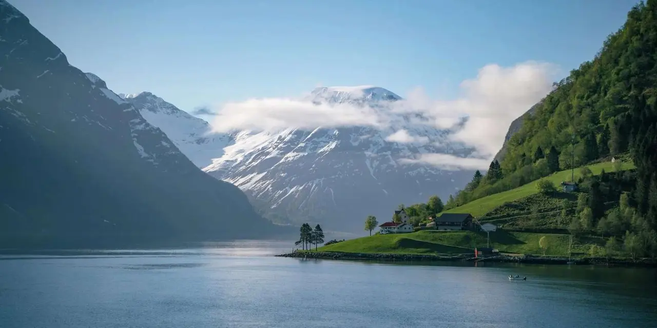 A calm fjord with snow-capped mountains behind and a green headland with small farm buildings at the water's edge