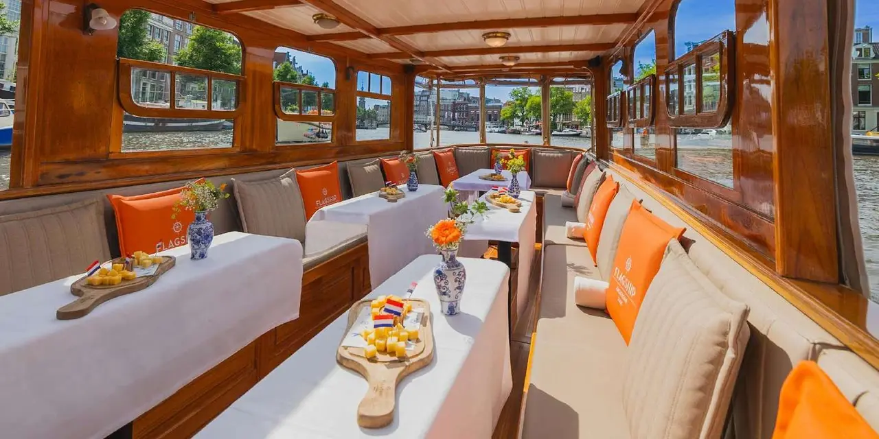 The inside of a wood-panelled saloon boat with white-clothed tables set with cheese boards, orange cushions and canal houses visible through the windows