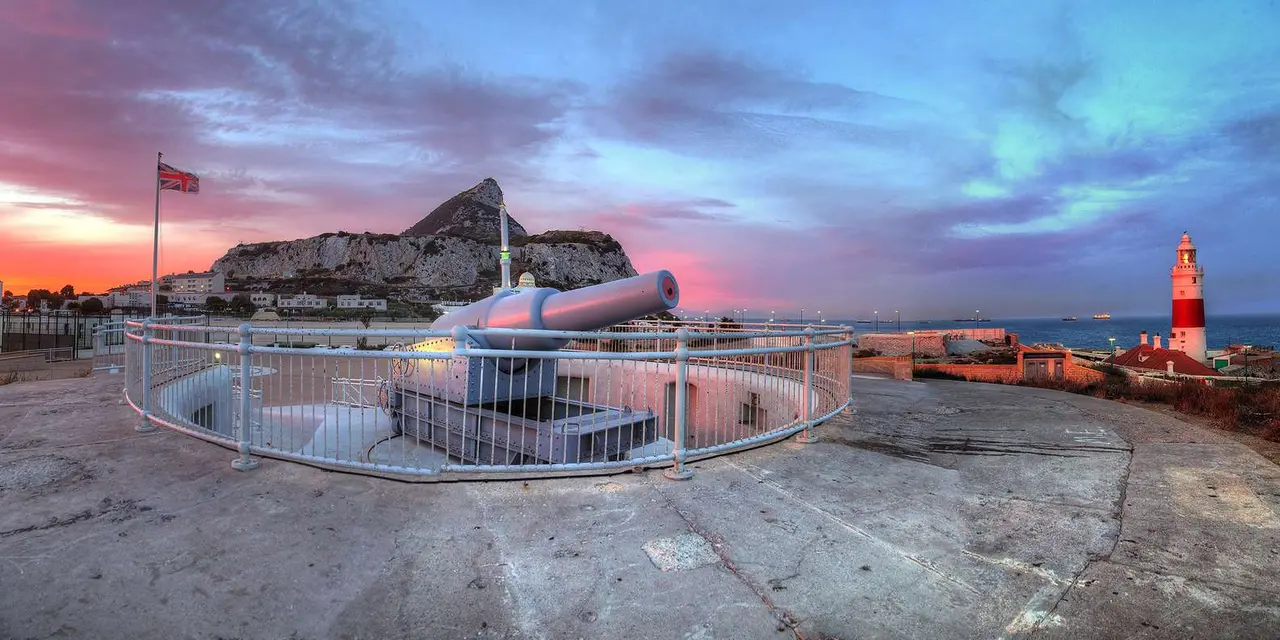 A large coastal gun on a round railed platform with the Rock of Gibraltar behind under a pink sunset sky