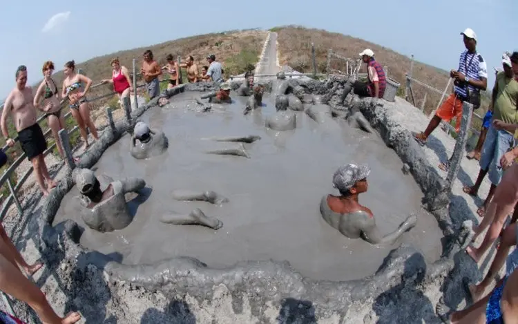 Bathers coated in grey mud inside the crater pool of a mud volcano, with people watching from the rim