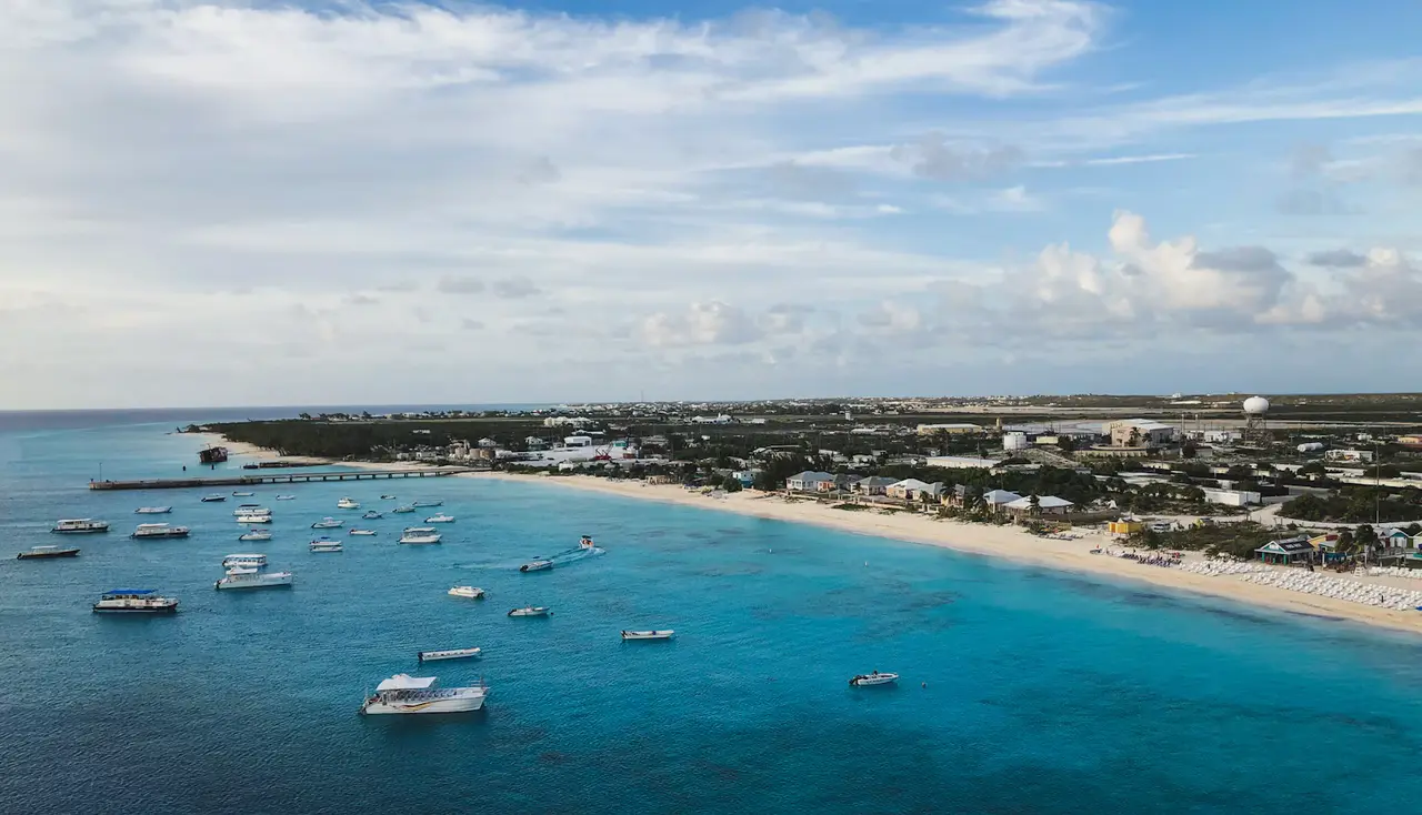 Aerial view of Grand Turk's west coast: small boats moored in clear turquoise water off a long white beach, with low-rise buildings behind and a wide cloud-streaked sky
