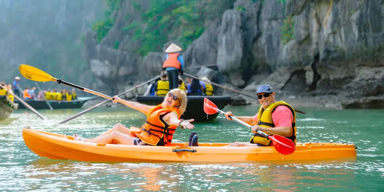 A couple in an orange kayak smiling, with a boatman in a conical hat poling a sampan behind them in a limestone cave lagoon
