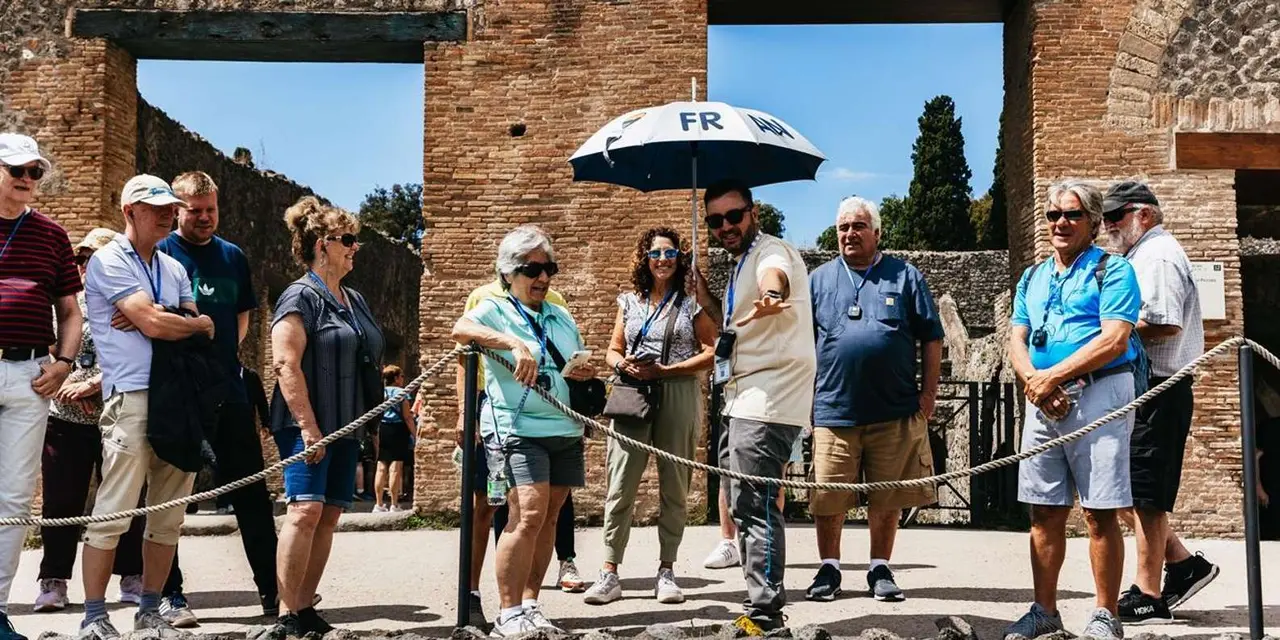 A tour group gathered around a guide holding an umbrella beside an ancient brick gateway in Pompeii