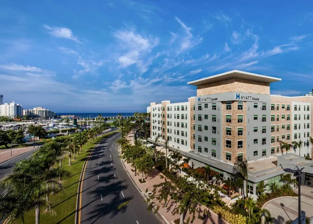 Hyatt House San Juan's beige tower beside a palm-lined boulevard, with the marina and the sea behind under a blue sky