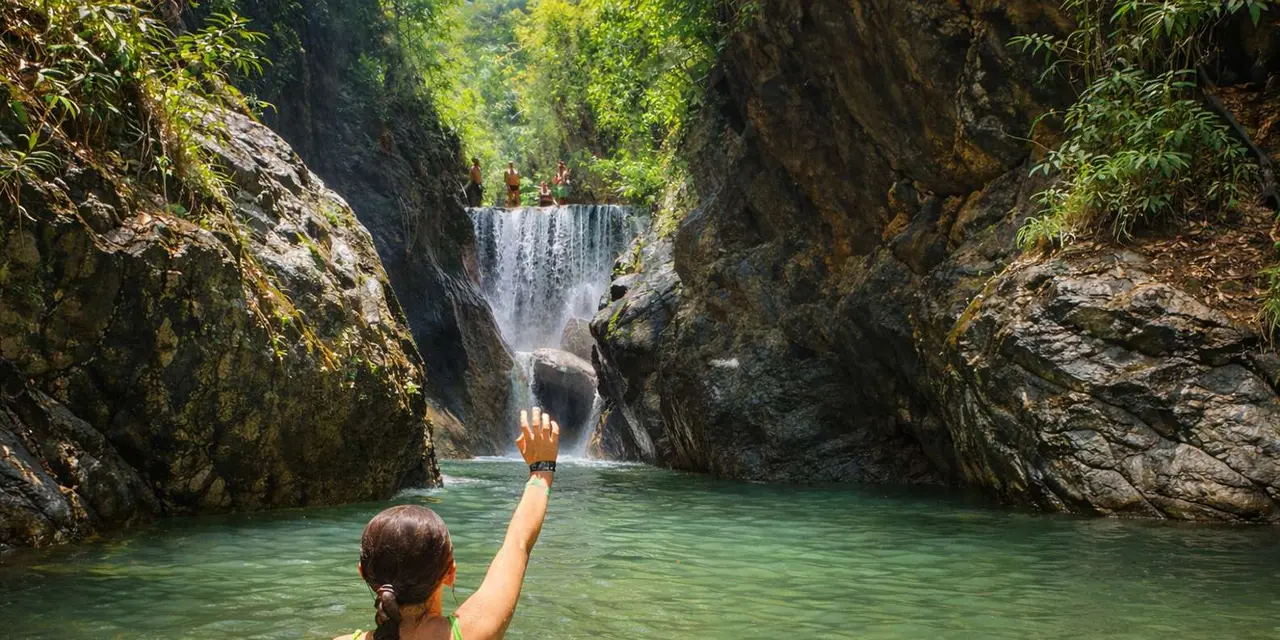 A woman standing in a green river pool with one arm raised toward a waterfall between two rock walls in a forested canyon