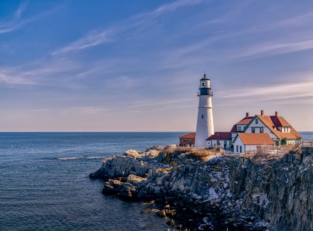 Portland Head Light, a white lighthouse beside a red-roofed keeper's house on a rocky cliff above a calm blue sea, under a streaked pale-blue and pink sky; the lighthouse is in Cape Elizabeth, about 3 miles from the cruise piers