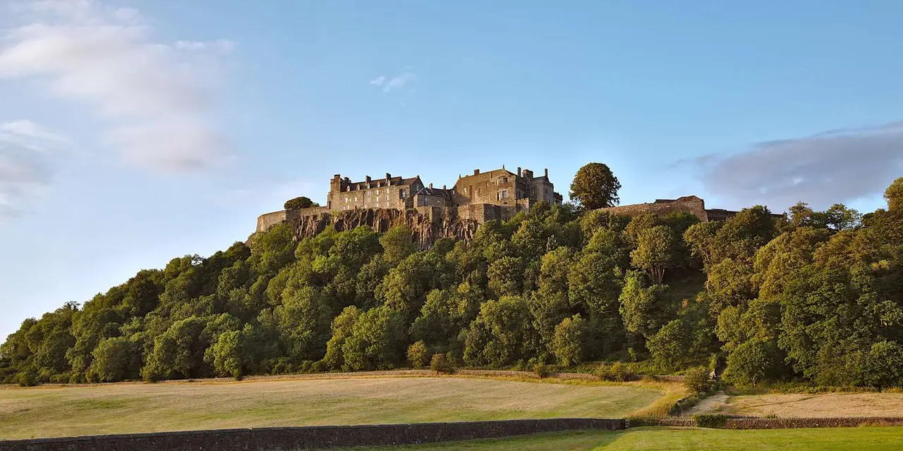 Stirling Castle's long stone walls on top of a wooded crag, with open fields below and a pale blue sky