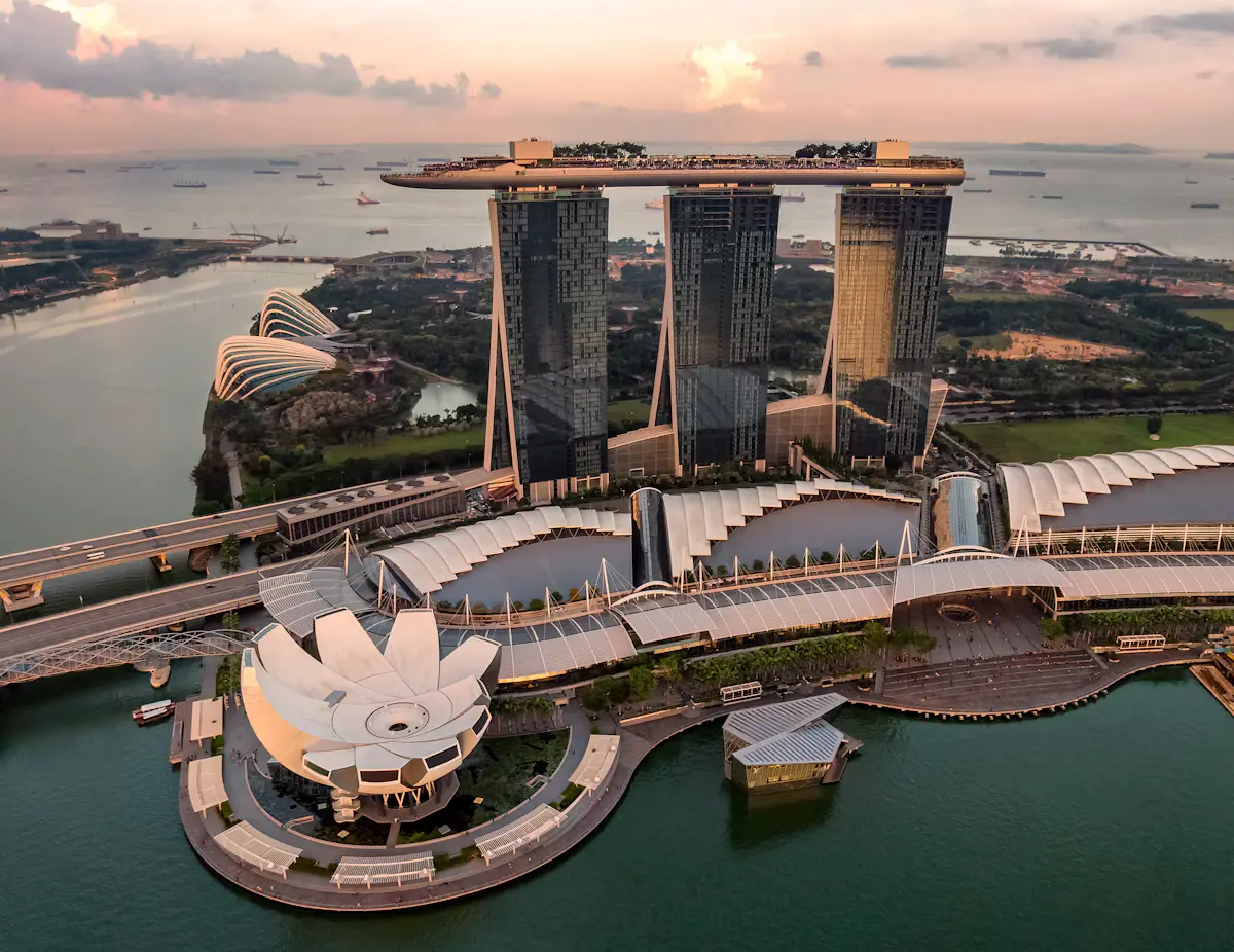 Aerial view of Marina Bay Sands' three towers and rooftop SkyPark at sunset, with the lotus-shaped ArtScience Museum on the water below and ships anchored offshore