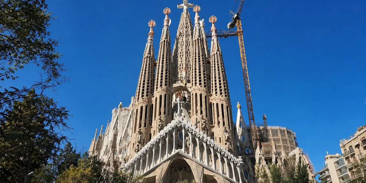 The towers and Passion facade of the Sagrada Família seen from the street, framed by trees against a deep blue sky with a construction crane beside it