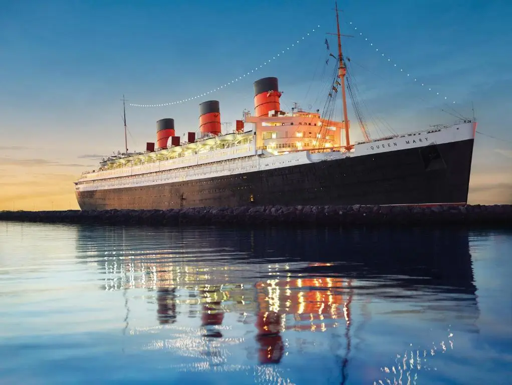 The Queen Mary ocean liner with its red and black funnels lit up at dusk on calm Long Beach water