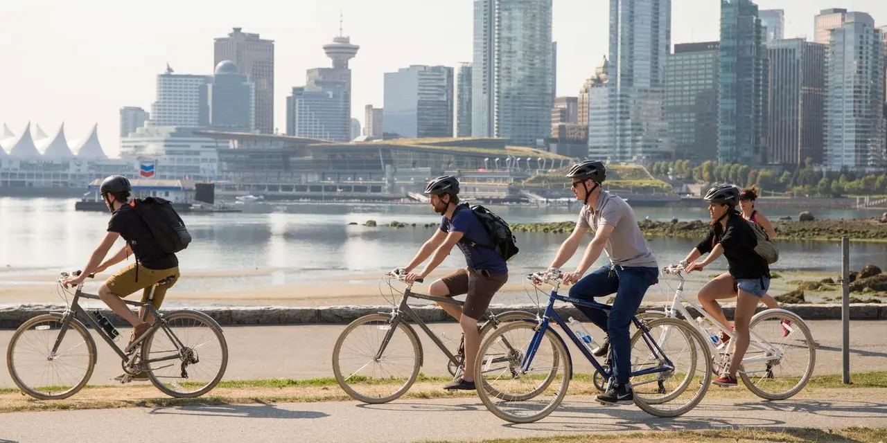 Four cyclists in helmets riding along the seawall, with Canada Place's white sails and the downtown towers across the water