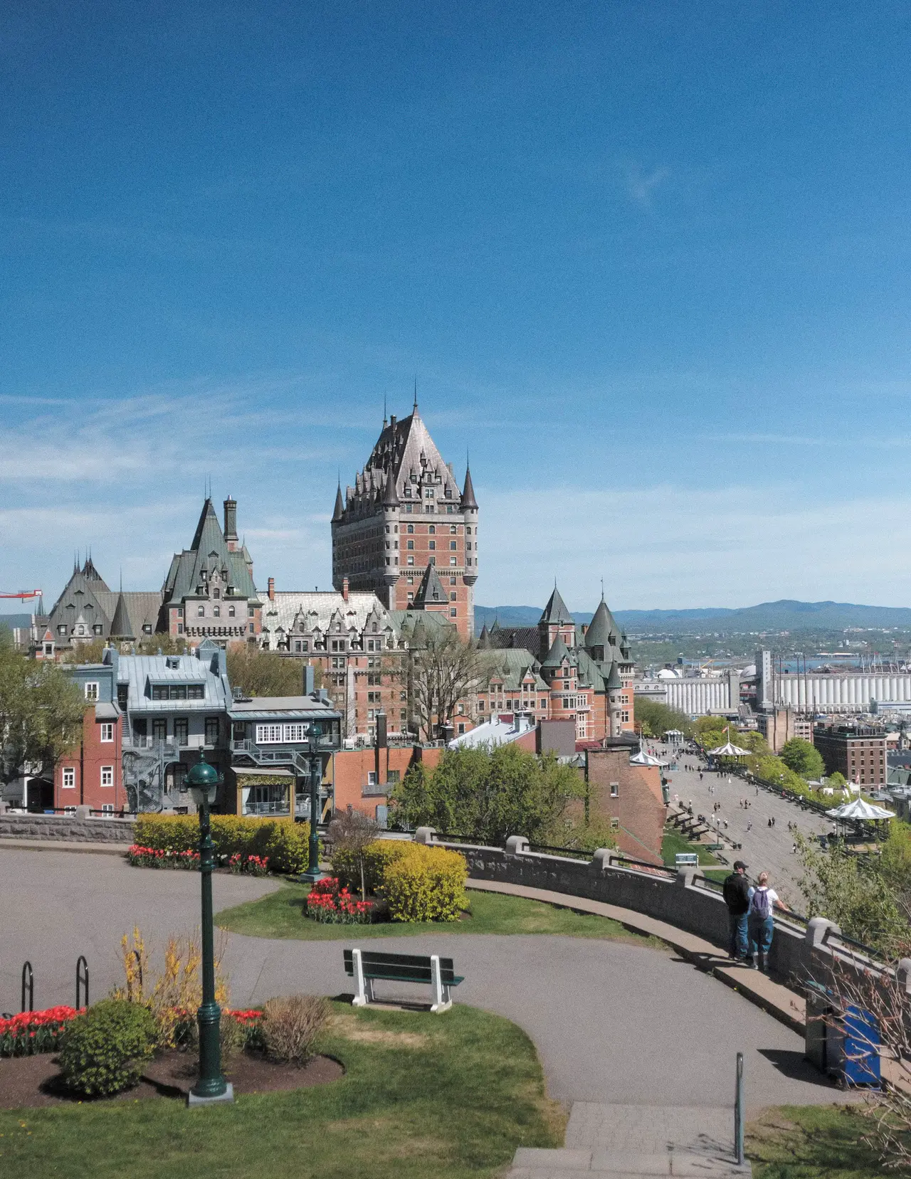 The Château Frontenac's green copper roofs and towers rising over Old Québec, seen from a garden path with a bench, under a wide clear blue sky