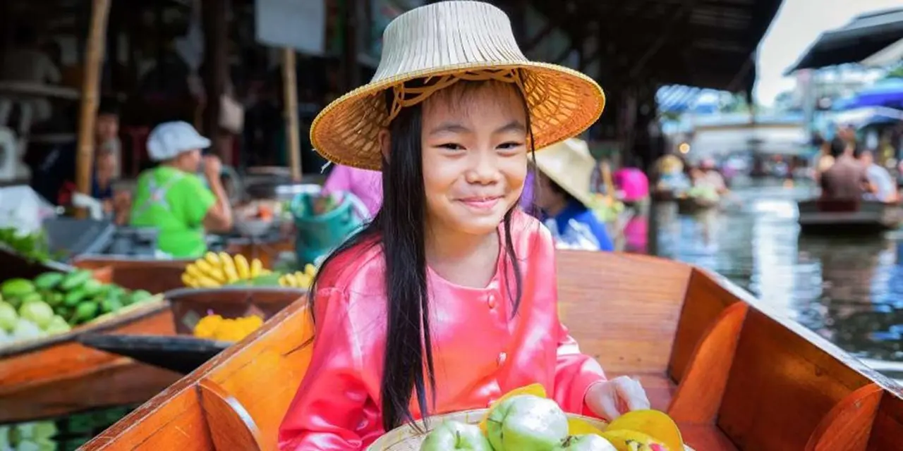 A smiling girl in a straw hat holding a basket of tropical fruit on a market boat