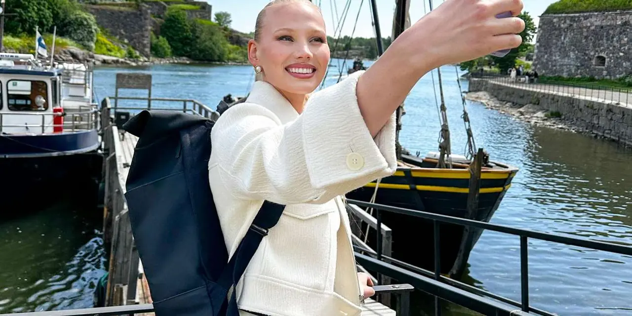 A traveller with a backpack takes a selfie on a harbour wall, with a moored wooden sailing ship and green rocky shore behind