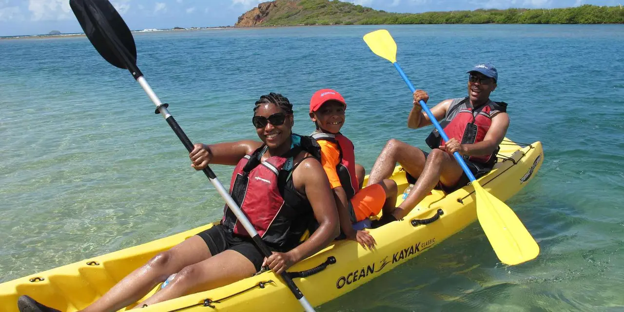 Three people in life vests paddling a yellow sit-on-top kayak across calm blue water, with a low green cay behind them