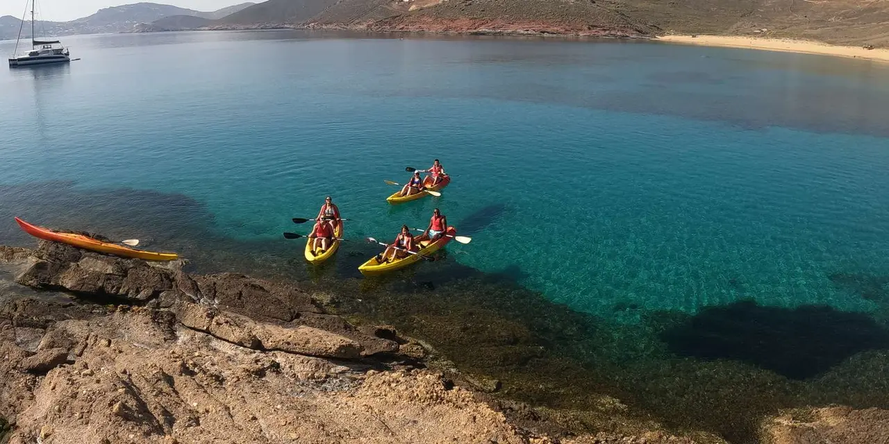 Three kayaks on clear turquoise water below a rocky shore, with a sailboat anchored in the bay