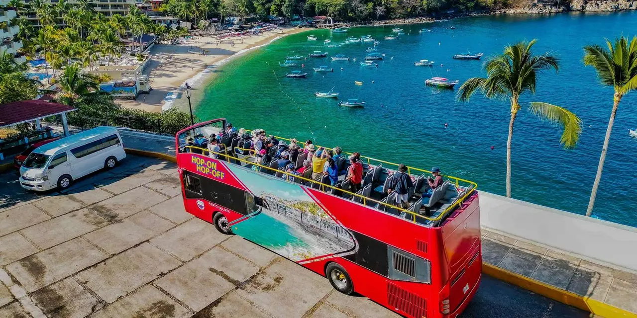 A red open-top hop-on-hop-off bus full of passengers parked above a green bay with a curved beach and moored boats