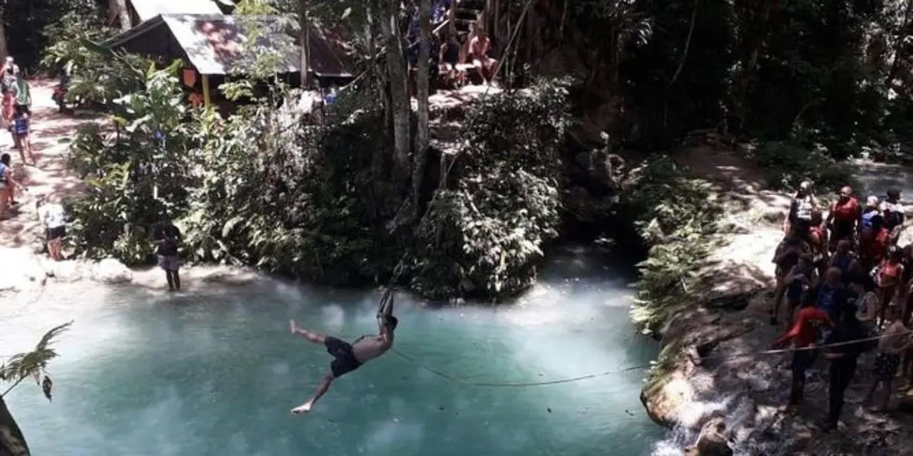 A swimmer mid-swing on a rope over a blue pool in the forest