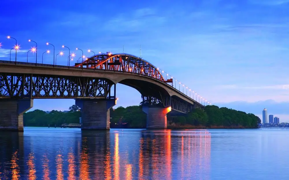 The Auckland Harbour Bridge lit at dusk, its lights reflected in the still water below