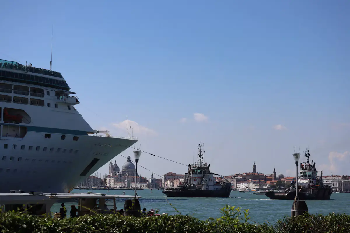 A white cruise ship moored on a Venice quay with two tugboats in the blue lagoon and the dome of Santa Maria della Salute on the skyline behind