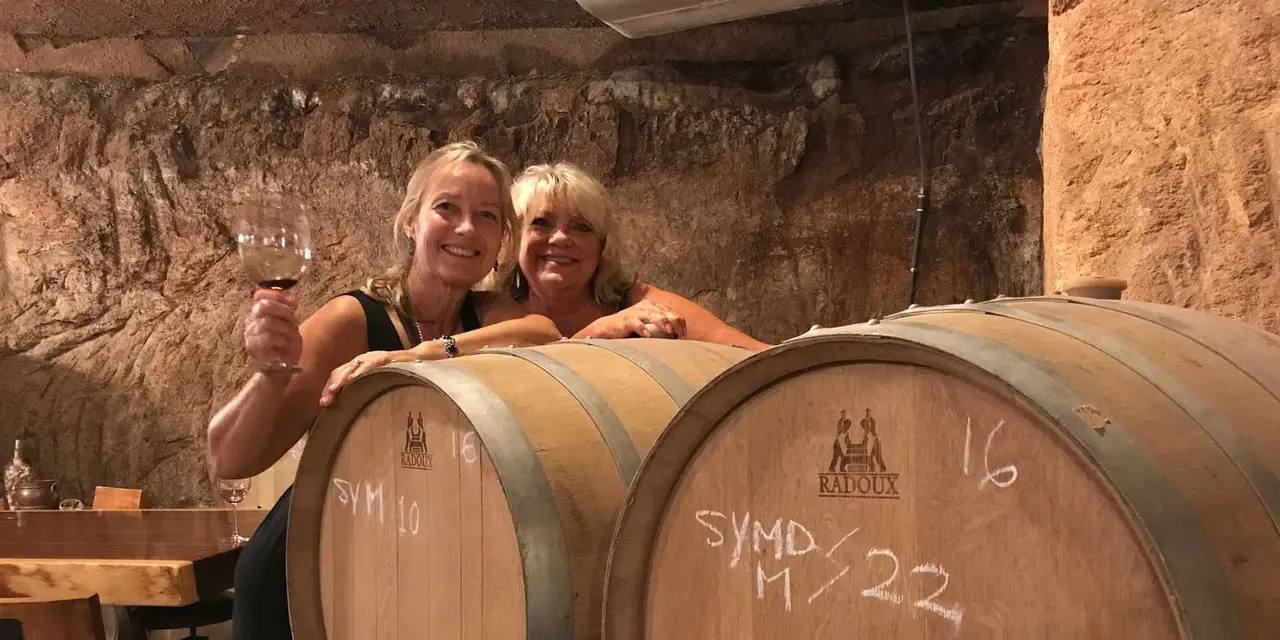 Two smiling women raising glasses of red wine beside oak barrels in a winery cellar