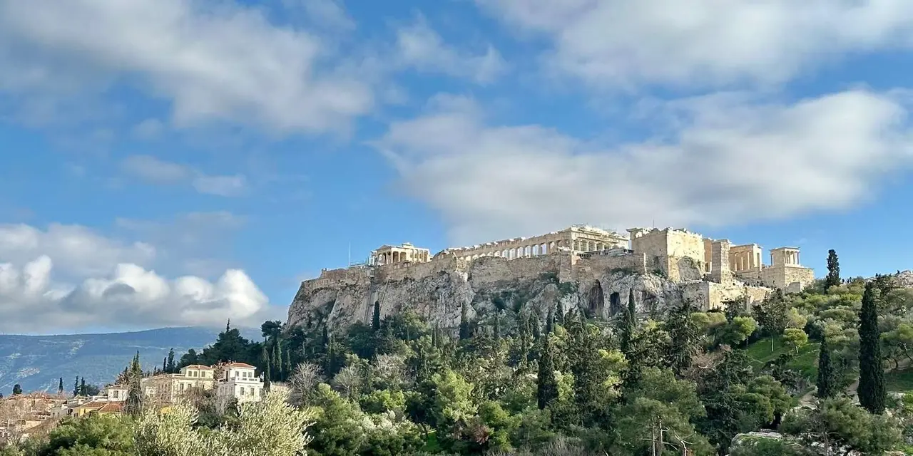 The Acropolis on its hill seen across green treetops under a cloudy blue sky
