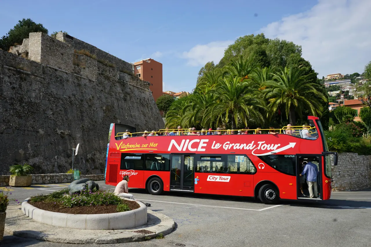 A red open-top Nice Le Grand Tour bus parked beside the stone walls of Villefranche's citadel