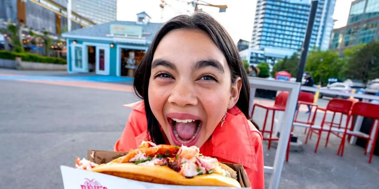A delighted woman holding up a lobster roll on the waterfront