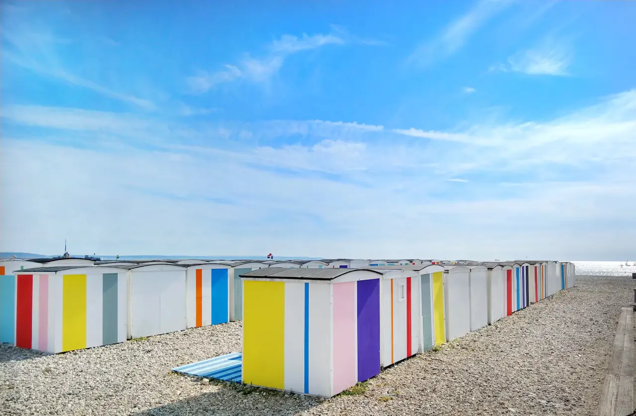 Rows of colourful striped beach huts on Le Havre's pebble beach under a bright blue sky, with the sea on the horizon
