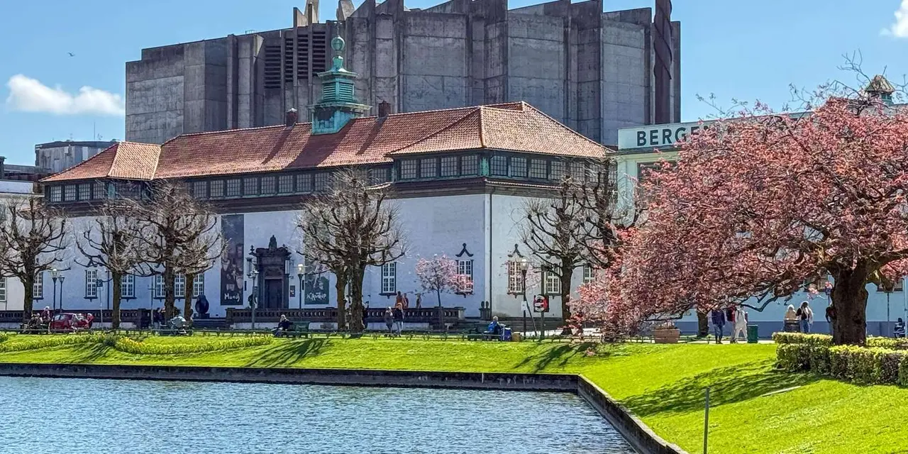 A large historic building with a green-topped tower beside a calm city lake and a manicured lawn