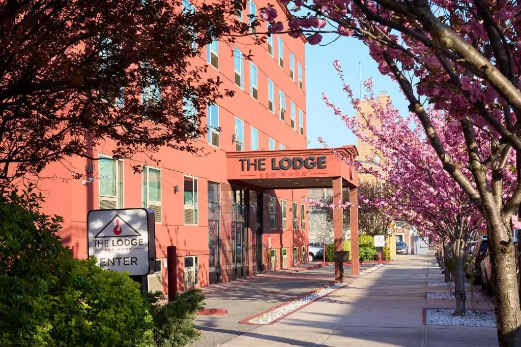 The red brick front of The Lodge Red Hook on a sunny day, its sign over the entrance and pink cherry blossoms along the sidewalk