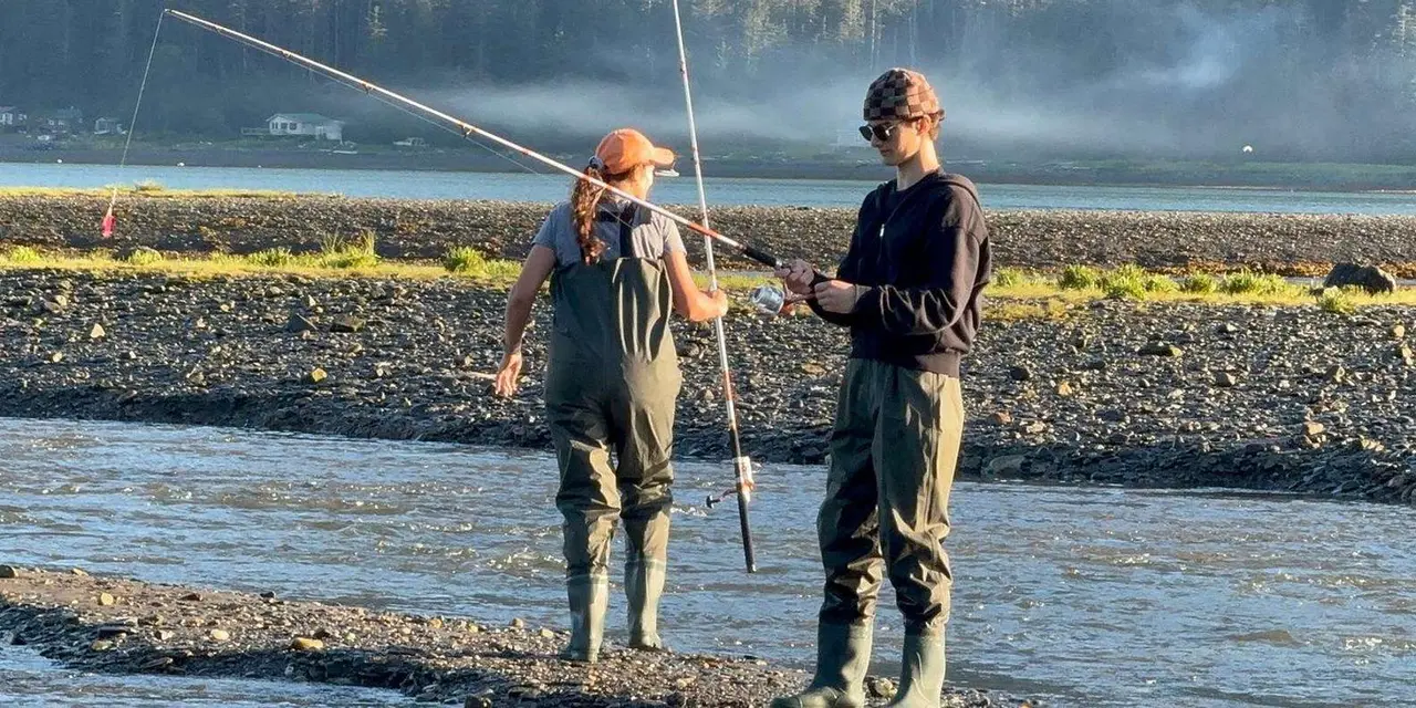 Two anglers in chest waders and boots holding fishing rods on a gravel bar beside a shallow stream, with a misty shoreline behind