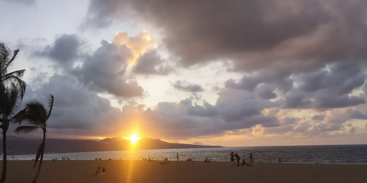 The sun setting through clouds over a sandy beach with a headland on the horizon