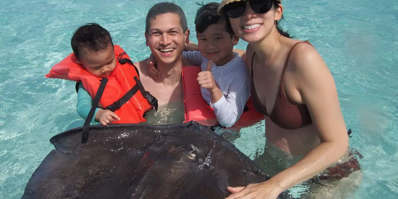 Two adults and two children in shallow turquoise water smiling over a large stingray, with boats anchored behind them