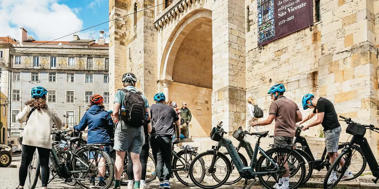 A group of cyclists in blue helmets gathered with e-bikes beside the pale stone arch of Lisbon's cathedral