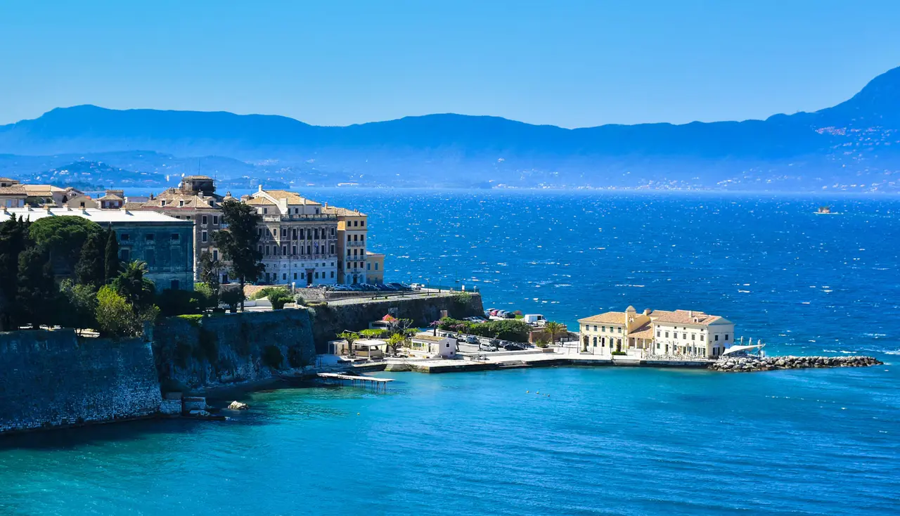 Corfu Town's seafront seen from the Old Fortress: old stone walls and pale stone buildings above bright turquoise water, with the mountains of mainland Greece across the strait