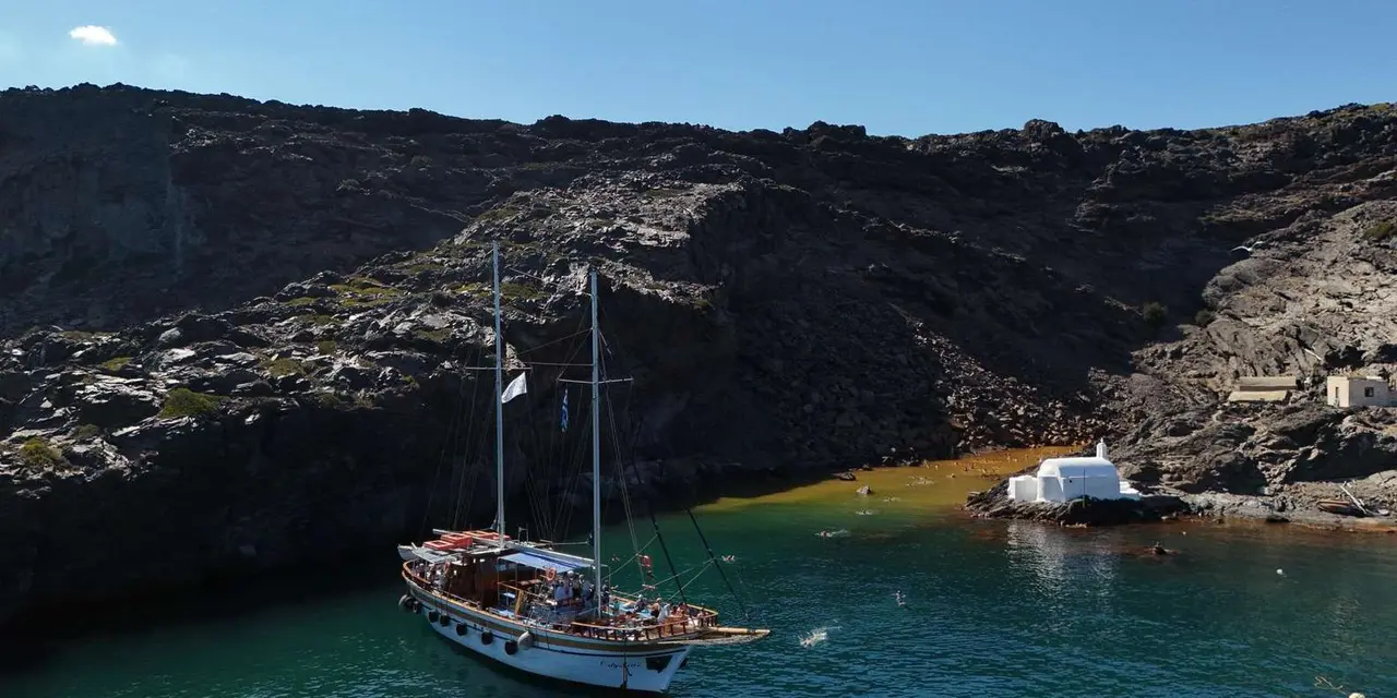A wooden two-masted boat anchored in a cove of black volcanic rock beside a small white chapel, with swimmers in the water